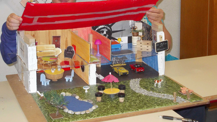 Photographie d’une maquette de maison comportant plusieurs pièces, un garage et un jardin. Un enfant soulève le toit en carton.