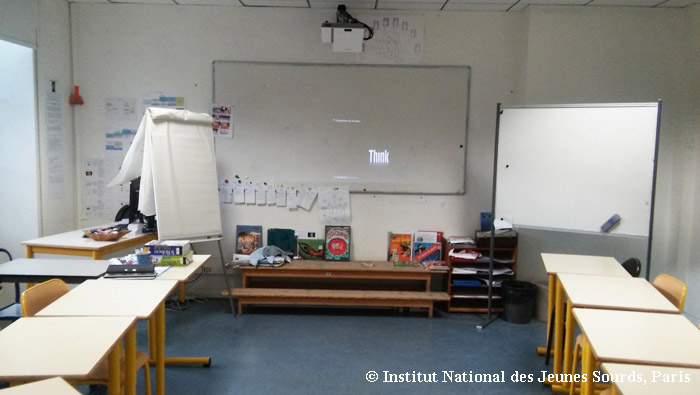 Photographie d’une salle de classe. Sur un mur, un tableau blanc surmonté d’un vidéoprojecteur. À droite du tableau principal : un tableau magnétique mobile ; à gauche : un paperboard.