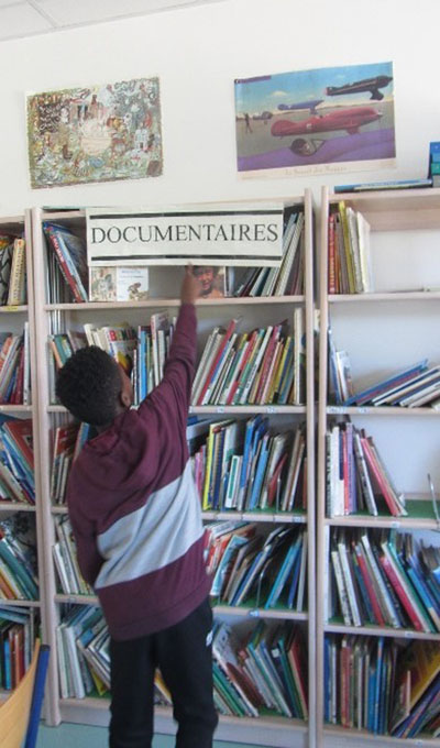 L’image est une photographie d’un élève devant une étagère de livres étiquetée « Documentaires »