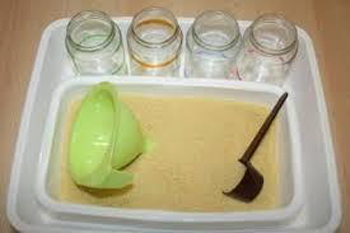 Photographie : une bassine contenant de la semoule. Des accessoires sont dans la bassine : une cuillère doseuse et un entonnoir. Quatre bocaux vides sans couvercles sont disposés derrière la bassine.