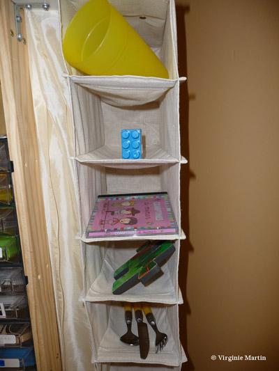 Photographie d'une étagère à cinq compartiments disposés verticalement. Dans les compartiments, de haut en bas : un gobelet, une brique de Lego, un CD, une pièce de puzzle, des couverts de table.