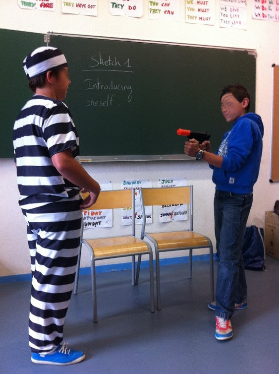 Photographie. Dans une classe d’école, deux élèves face à face, l’un est habillé d’un costume de prisonnier (blanc à rayures noires horizontales), l’autre le tient en joue avec un fusil jouet.